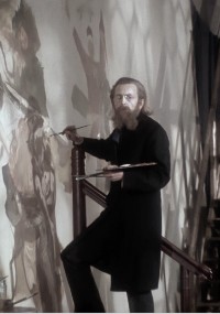 Mężczyzna z długimi włosami i brodą, ubrany w ciemne ubranie, stoi na schodach i maluje duży mural na ścianie, trzymając paletę i pędzel.