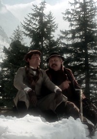 Dwóch mężczyzn w ubraniach vintage siedzi na zaśnieżonym terenie, na tle wiecznie zielonych drzew i gór, wydaje się, że rozmawiają i cieszą się zimowym krajobrazem.