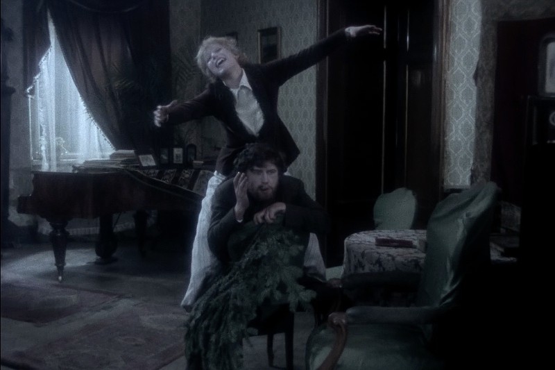 Kobieta stoi za siedzącym mężczyzną, pochyla się do przodu z wyciągniętymi ramionami i radosnym wyrazem twarzy. Mężczyzna siedzi na krześle, okrytym zielenią, w słabo oświetlonym, staromodnym salonie.