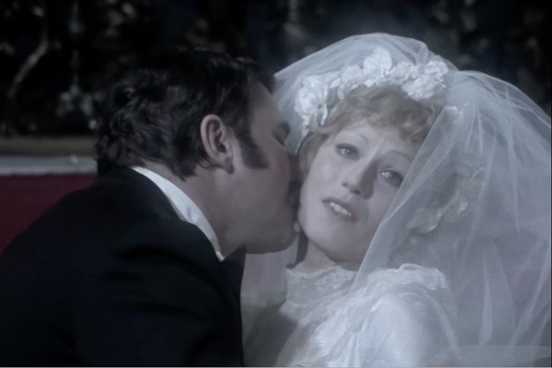 Pan młody całuje pannę młodą w policzek. Panna młoda, ubrana w białą suknię i welon z kwiatowym nakryciem głowy, patrzy w dal z odległym wyrazem twarzy. Tło jest słabo oświetlone.