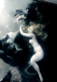 Rozmyty, eteryczny obraz nagiej osoby z długimi włosami, przyczepionymi do szyi ciemnego, groźnego konia lub zwierzęcia, tworzący surrealistyczną i dramatyczną atmosferę podwodną lub oniryczną.