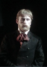 Mężczyzna o jasnych włosach i brodzie, ubrany w ciemny garnitur i dużą, bordową muszkę, stoi na ciemnym tle i wygląda poważnie.