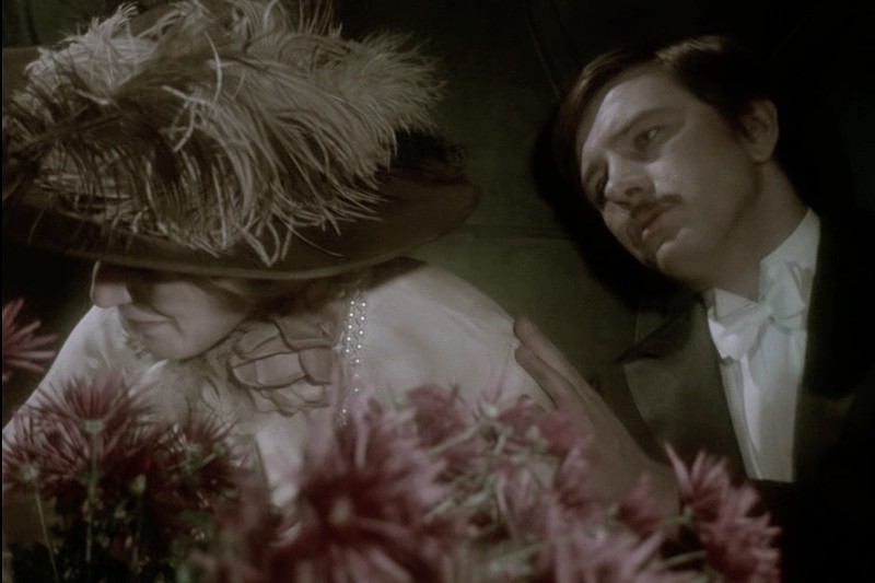 Mężczyzna w formalnym stroju delikatnie dotyka ramienia kobiety w dużym kapeluszu z piórami i perłami, otoczonej różowymi kwiatami. Scena ma atmosferę vintage, słabo oświetloną.