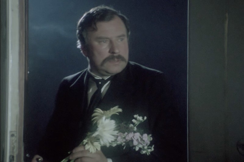 Mężczyzna z wąsami, ubrany w garnitur i krawat, stoi w drzwiach, trzymając mały bukiet kwiatów, patrząc w lewo z poważnym wyrazem twarzy. Tło jest słabo oświetlone.