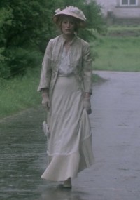 Kobieta w eleganckiej, długiej, jasnej sukni i kapeluszu idzie samotnie mokrą ścieżką, otoczona zielenią i mgłą, w staroświeckim, zabytkowym otoczeniu.