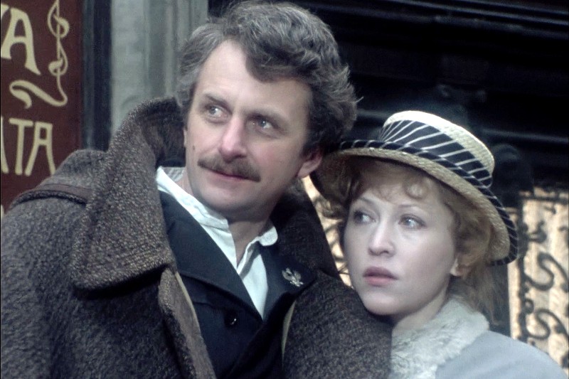 Mężczyzna z kręconymi włosami i wąsami, ubrany w gruby brązowy płaszcz, stoi obok kobiety w pasiastym kapeluszu i lekkim płaszczu. Oboje patrzą lekko w lewo z poważnymi minami, stojąc blisko siebie.