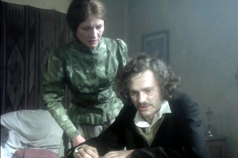 Kobieta w zielonej sukni z epoki wiktoriańskiej stoi obok siedzącego mężczyzny z kręconymi włosami i wąsami, który najwyraźniej pisze przy biurku w słabo oświetlonym, staromodnym pokoju.