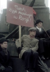 Trzech mężczyzn siedzi na kamiennych schodach; jeden trzyma czerwony znak z białym polskim tekstem, na którym widnieje napis Niech żyje rewolucja w Rosji!, co oznacza Niech żyje rewolucja w Rosji! Za nimi częściowo widać posąg.
