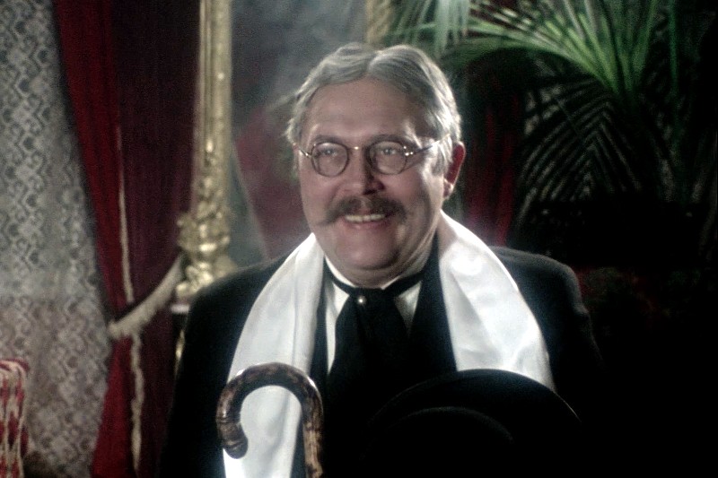 Starszy mężczyzna w okrągłych okularach, z wąsami i siwymi włosami uśmiecha się, mając na sobie biały szalik i czarny garnitur. Trzyma laskę i kapelusz, a w tle ozdobne zasłony i duża roślina.