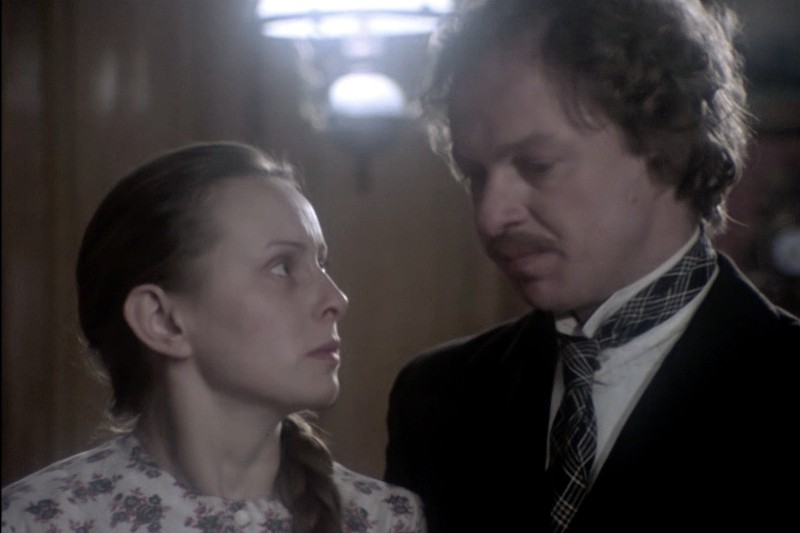 Kobieta w kwiecistej bluzce uważnie przygląda się mężczyźnie w garniturze i krawacie; stoją blisko siebie w pomieszczeniu, w łagodnym oświetleniu, w tle widać rozmazane światło lampy.