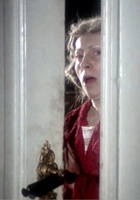 Kobieta w czerwonej szacie wygląda na zszokowaną lub przestraszoną, patrząc przez częściowo uchylone białe drzwi z ozdobną mosiężną klamką.