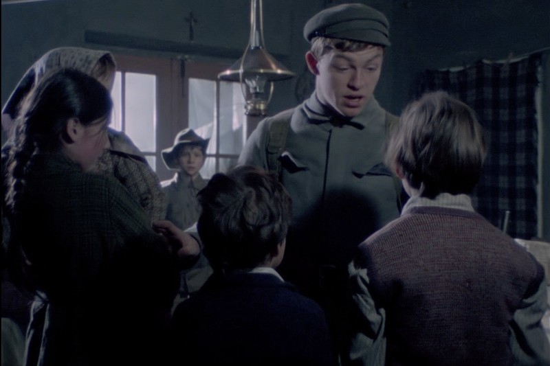 Młody żołnierz w mundurze stoi w słabo oświetlonym pomieszczeniu, otoczony dziećmi i kobietą; nad nim wisi lampa, a światło wpada przez okno w tle.