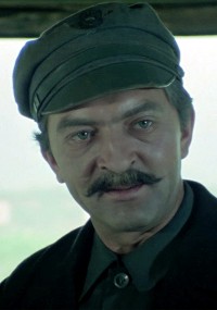 Mężczyzna o jasnych oczach i wąsach nosi ciemną czapkę i kurtkę, patrząc lekko na bok. Tło jest rozmyte i jasne.