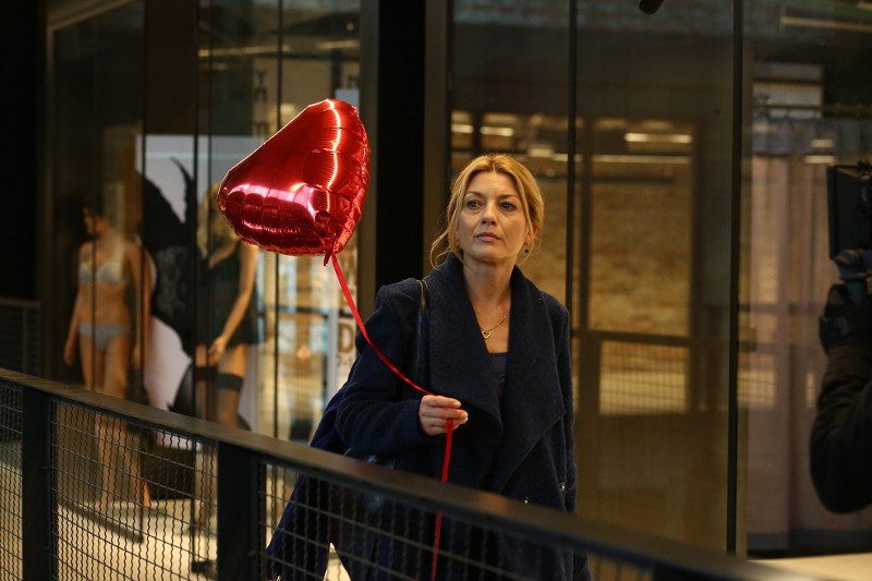 Kobieta w ciemnym płaszczu trzyma czerwony balon w kształcie serca, idąc w środku. W tle widać duże okna i reklamę z manekinami w bieliźnie.