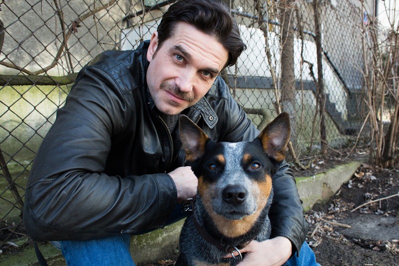 Mężczyzna w czarnej skórzanej kurtce kuca obok psa rasy blue heeler. Są na zewnątrz, w pobliżu ogrodzenia z siatki łańcuchowej i bezlistnych roślin, obaj patrzą prosto w kamerę.