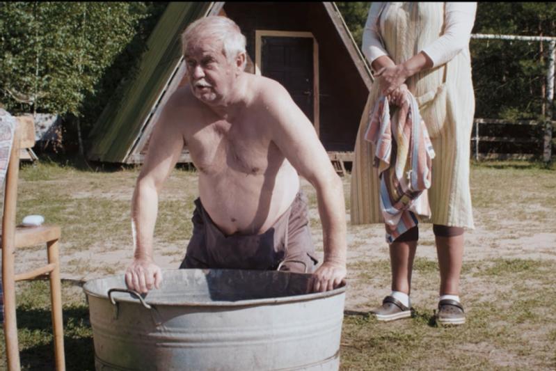 Starszy mężczyzna bez koszuli pochyla się nad dużą metalową wanną na zewnątrz, podczas gdy kobieta stojąca w pobliżu trzyma ręcznik. W tle widać drzewa i chatę.