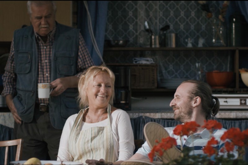 Trzy osoby w przytulnej kuchni: starszy mężczyzna stoi trzymając kubek, podczas gdy uśmiechnięta kobieta w fartuchu i młodszy mężczyzna z brodą siedzą przy stole i rozmawiają. Na pierwszym planie znajdują się czerwone kwiaty.