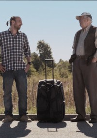 Dwóch mężczyzn stoi na drodze obok dużej czarnej walizki. Jeden z nich ma na sobie kraciastą koszulę i dżinsy z kucykiem, a drugi jest starszy, ma na sobie kapelusz, kamizelkę i spodnie khaki. W tle widać drzewa i błękitne niebo.