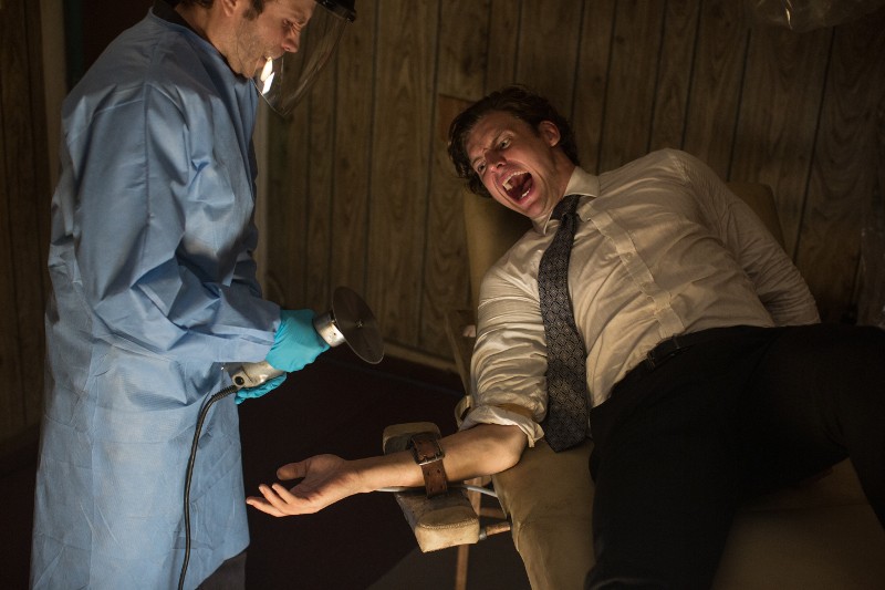 Mężczyzna w garniturze krzyczy ze strachu, przywiązany do krzesła, podczas gdy inna osoba w stroju ochronnym trzyma elektronarzędzie w pobliżu jego odsłoniętej ręki w ciemnym pomieszczeniu o drewnianych ścianach.