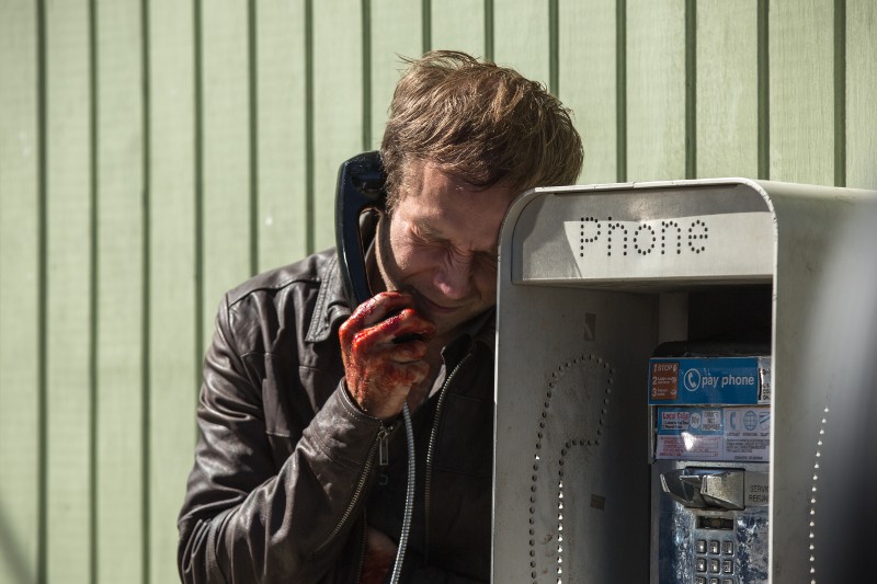 Zrozpaczony mężczyzna z krwią na dłoni opiera się o automat telefoniczny, trzymając słuchawkę przy uchu, z zamkniętymi oczami i napiętą twarzą. W tle widać jasnozieloną ścianę panelową.