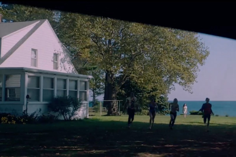 Cztery osoby biegną przez trawiaste podwórko w kierunku białego domu z zadaszonymi gankami, dużymi drzewami i widokiem na błękitne jezioro w tle pod czystym niebem.