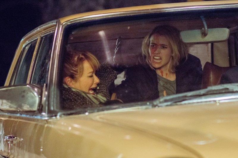 Dwie kobiety siedzą na przednich siedzeniach jasnobrązowego samochodu nocą, obie wyglądają na zdenerwowane. Kobieta po lewej krzyczy, podczas gdy kobieta po prawej wygląda na przerażoną z krwią na twarzy.