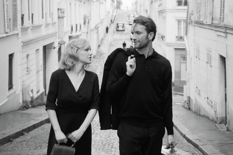 Kobieta i mężczyzna idą obok siebie po brukowanej ulicy, niosąc walizki. Oboje mają na sobie ciemne ubrania i patrzą na siebie, w tle widać budynki i stromą ulicę. Obraz jest czarno-biały.