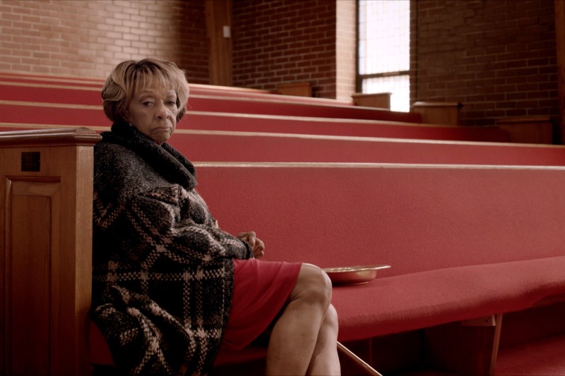 Starsza kobieta w kraciastym płaszczu siedzi sama na czerwonych ławkach w pustym kościele, wyglądając na zamyśloną. Taca na datki leży obok niej. Światło słoneczne sączy się przez wysokie okno, oświetlając ceglane ściany.