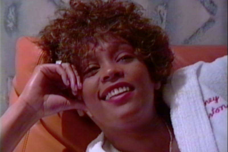 Kobieta o kręconych brązowych włosach uśmiecha się, wypoczywając na pomarańczowej powierzchni, ubrana w białą szatę i opierając głowę na ręce.