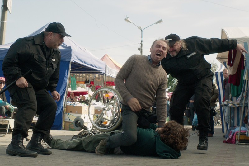 Pewien mężczyzna wściekle atakuje inną osobę leżącą na ziemi obok przewróconego wózka inwalidzkiego, podczas gdy dwóch umundurowanych funkcjonariuszy próbuje interweniować na targu pod gołym niebem.