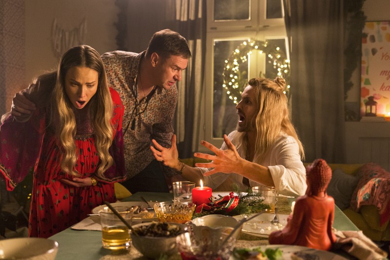 Trzy osoby siedzą przy świątecznym stole, reagując szokiem i zaskoczeniem. Stół jest zastawiony jedzeniem, świecami i świątecznymi dekoracjami, z ciepłym oświetleniem i udekorowanym oknem w tle.