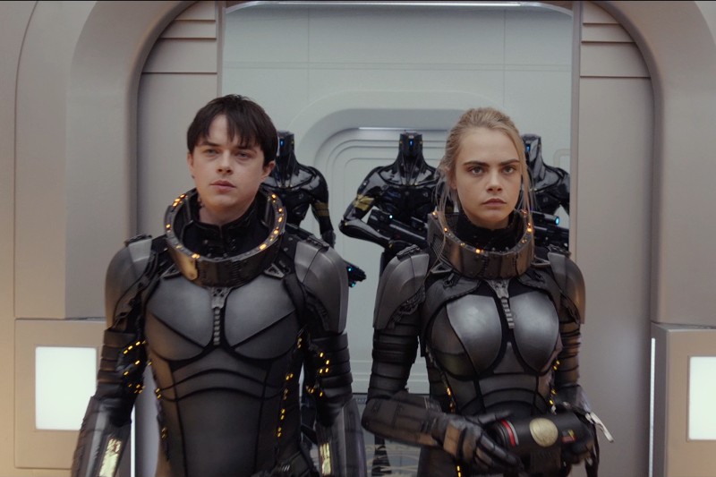 Dwie osoby w futurystycznych, opancerzonych skafandrach kosmicznych idą obok siebie w korytarzu high-tech, za nimi w tle podążają roboty. Sceneria i ich miny sugerują poważną lub napiętą sytuację.