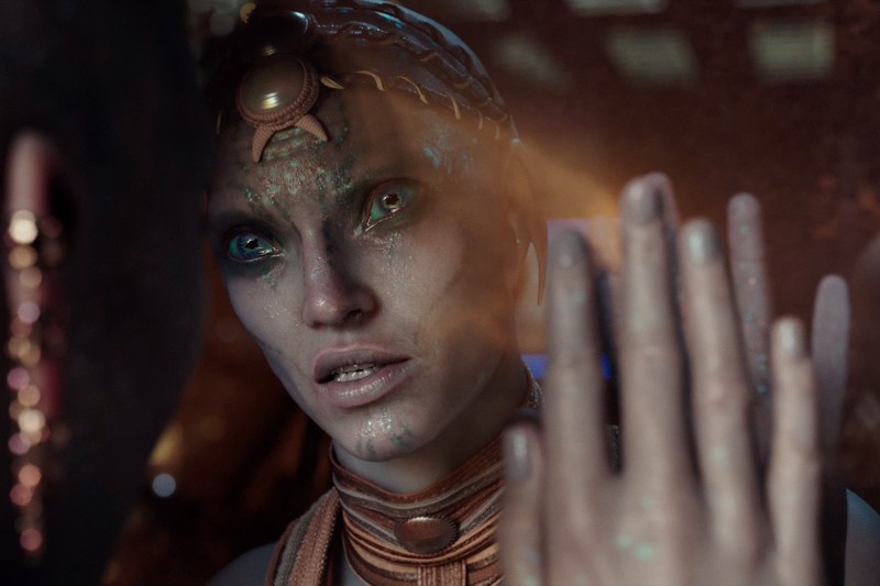 Humanoidalna postać o opalizującej, gadziej skórze, świecących zielonych oczach i ozdobnej biżuterii dotyka odbijającej powierzchni, wpatrując się uważnie z mieszanką ciekawości i troski. Ciepłe światło oświetla jej twarz i dłoń.