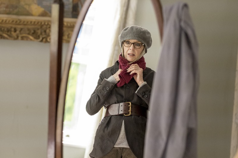 Kobieta w okularach, szarym berecie i grubym brązowym pasku poprawia czerwony szalik, patrząc w wysokie lustro w dobrze oświetlonym pokoju.