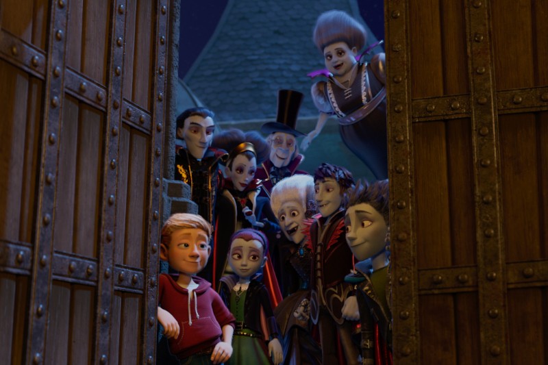 Grupa animowanych postaci, w tym blond chłopiec i różni wampiry w kolorowych strojach, zbierają się blisko siebie i zaglądają przez duże, otwarte drewniane drzwi, sprawiając wrażenie ciekawych i podekscytowanych.