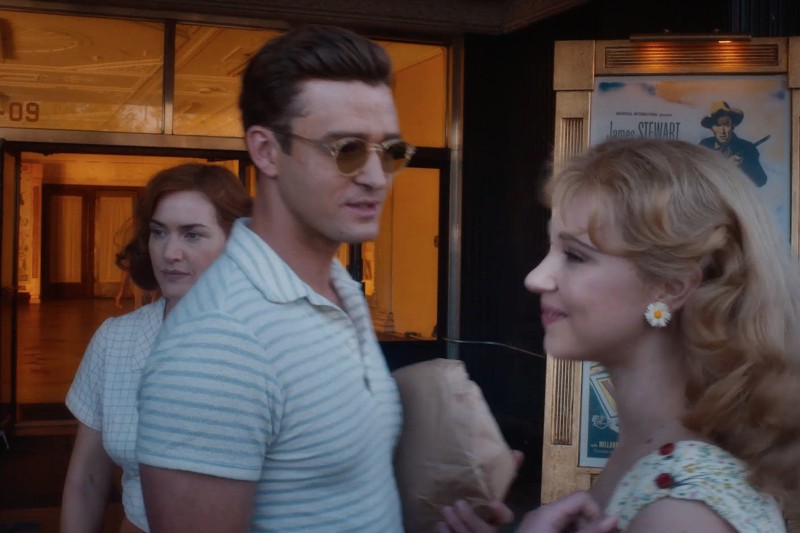 Mężczyzna w okularach przeciwsłonecznych trzymający papierową torbę stoi między dwiema kobietami ubranymi w ubrania vintage, na zewnątrz budynku, w tle którego wiszą plakaty filmowe.