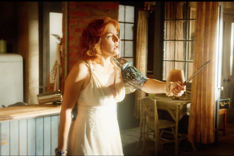 Kobieta z rudymi włosami w białej sukience stoi w oświetlonym słońcem pokoju, trzymając przed sobą nóż w geście obronnym. W tle znajdują się rustykalne meble, lodówka i ceglane ściany.