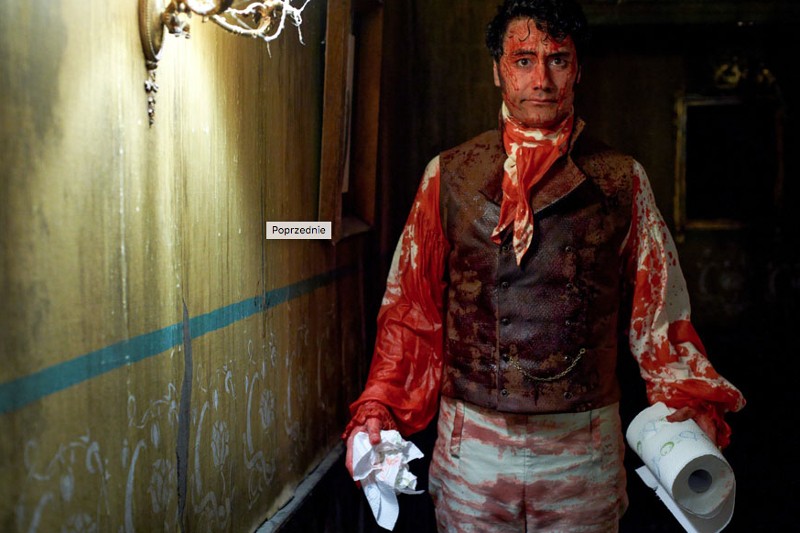 Mężczyzna w stroju w stylu wiktoriańskim zachlapanym krwią trzyma rolkę papieru toaletowego i chusteczki higieniczne, stojąc w słabo oświetlonym, staromodnym pokoju z łuszczącą się tapetą i lampą ścienną.