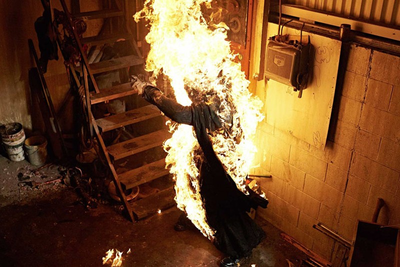 Osoba ubrana w ciemny płaszcz stoi w płomieniach przy drewnianych schodach w słabo oświetlonym pomieszczeniu przemysłowym, z wyciągniętymi przed siebie ramionami i ciałem objętym ogniem.