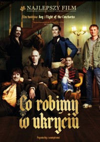 Pięciu mężczyzn w różnych strojach i pozach stoi i siedzi w słabo oświetlonym, staromodnym pokoju z zamontowanymi porożami na ścianie. Na dole widnieje tytuł polskiego filmu Co robimy w ukryciu.
