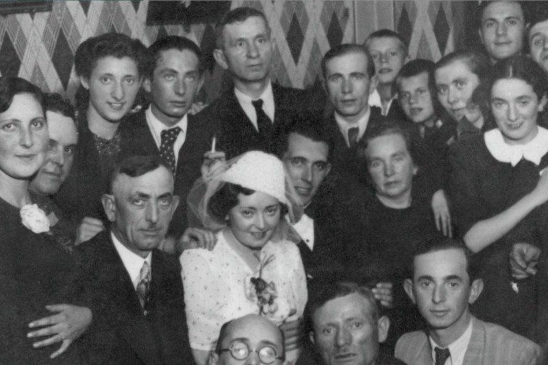 Czarno-białe zdjęcie dużej grupy ludzi, w tym panny młodej w białej sukni i kapeluszu, w otoczeniu rodziny i przyjaciół, pozujących blisko siebie w pomieszczeniu na tle wzorzystej ściany.