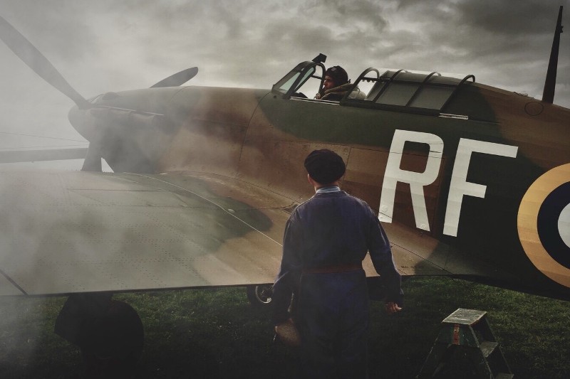 Osoba w kombinezonie ochronnym stoi obok zabytkowego samolotu wojskowego oznaczonego RF, podczas gdy pilot siedzi w kokpicie, otoczony mgłą na trawiastym polu.