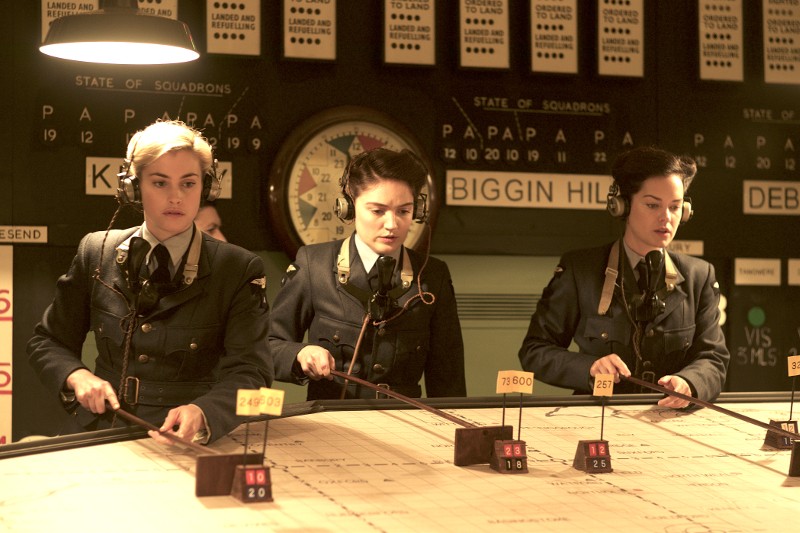 Trzy kobiety w mundurach z czasów II wojny światowej i z zestawami słuchawkowymi używają wskaźników na dużej mapie w sali operacyjnej, prawdopodobnie koordynując misje lotnicze. Sceneria ma charakter vintage z zegarami i tablicami informacyjnymi w tle.