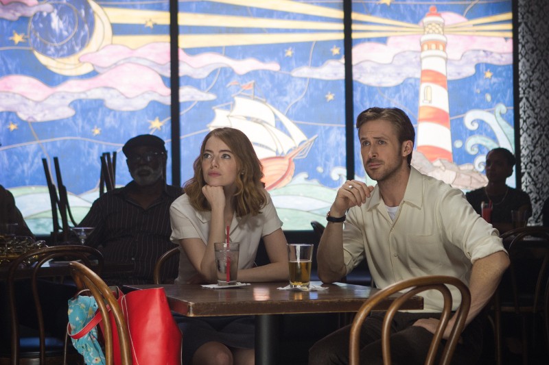 Kobieta i mężczyzna siedzą przy stoliku w kawiarni, zamyśleni patrząc przed siebie. Mają przed sobą drinki. Za nimi znajduje się kolorowy mural przedstawiający statek, latarnię morską, chmury i gwiazdy.