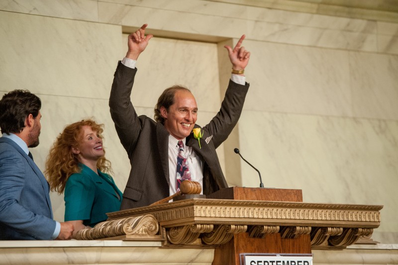 Mężczyzna w garniturze stoi na podium, uśmiecha się i triumfalnie podnosi obie ręce. Dwie osoby obok niego, mężczyzna i kobieta, patrzą z uśmiechami. Sceneria wydaje się być formalną salą rządową lub sądową.