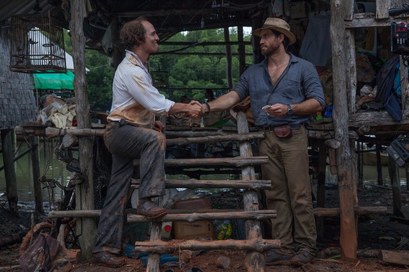 Dwóch mężczyzn, jeden stojący na drewnianych schodach, a drugi na ziemi, ściska sobie dłonie na zewnątrz rustykalnej drewnianej chaty. Obaj mają na sobie solidne ubrania i kapelusze; w tle widać błotnistą ziemię i zielone liście.