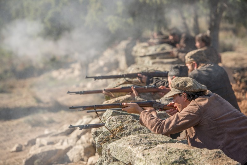 Kilka osób ubranych w staromodne stroje kuca za kamiennym murem, celując i strzelając w zadymione pole bitwy, co sugeruje historyczną bitwę lub scenę rekonstrukcji bitwy rozgrywającą się na świeżym powietrzu, w wiejskim otoczeniu.