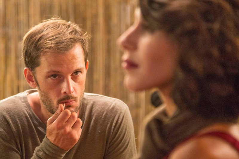 Mężczyzna o zamyślonym wyrazie twarzy opiera dłoń na brodzie, wpatrując się w kobietę na pierwszym planie, która jest nieostra. Tło jest delikatnie oświetlone złotymi tonami.