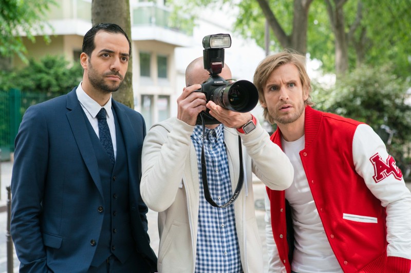 Trzech mężczyzn stoi na zewnątrz; jeden trzyma aparat i robi zdjęcie. Mężczyzna po lewej nosi garnitur, a mężczyzna po prawej czerwono-białą kurtkę uniwersytecką. Wszyscy wydają się być skupieni na czymś przed sobą.