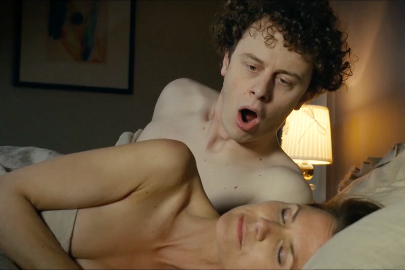 Mężczyzna o kręconych włosach wygląda na zaskoczonego, siedząc na łóżku obok kobiety, która leży z zamkniętymi oczami, częściowo przykryta prześcieradłem. W tle widać zapaloną lampę.
